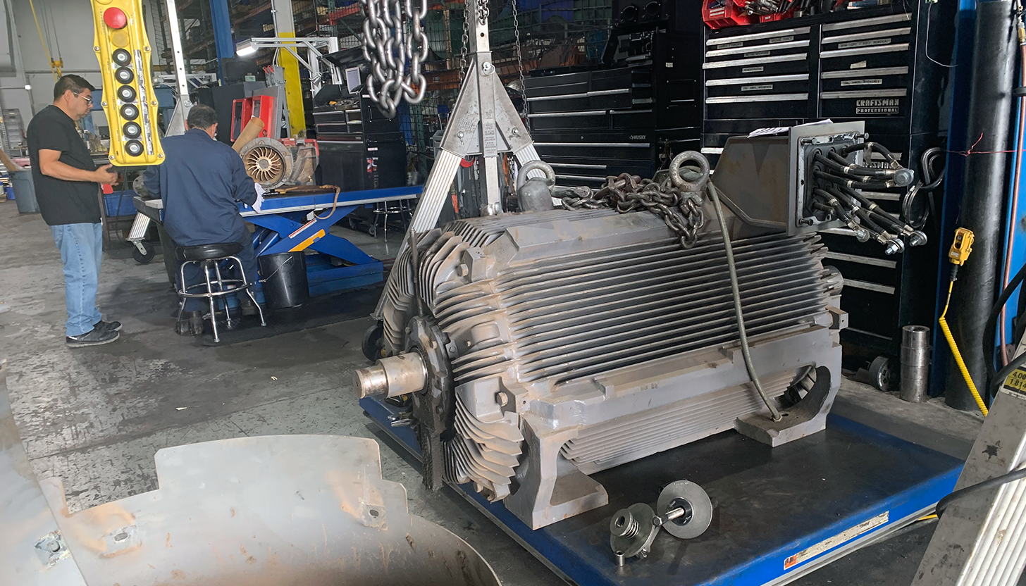 Large AC/DC electric motor on a hydraulic table under a 2-ton gantry crane at Apex Industrial Automation, with two technicians discussing repair strategies in the background.