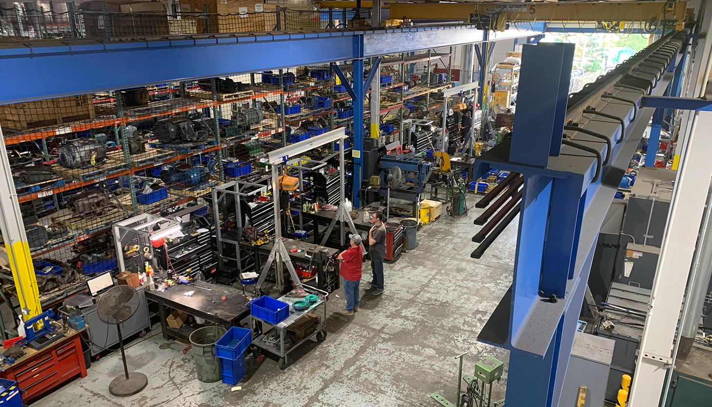 Overhead view of Apex Industrial Automation's repair shop featuring numerous workbenches with mobile gantry cranes, an overhead 15-ton gantry crane, and a stock of large motors.