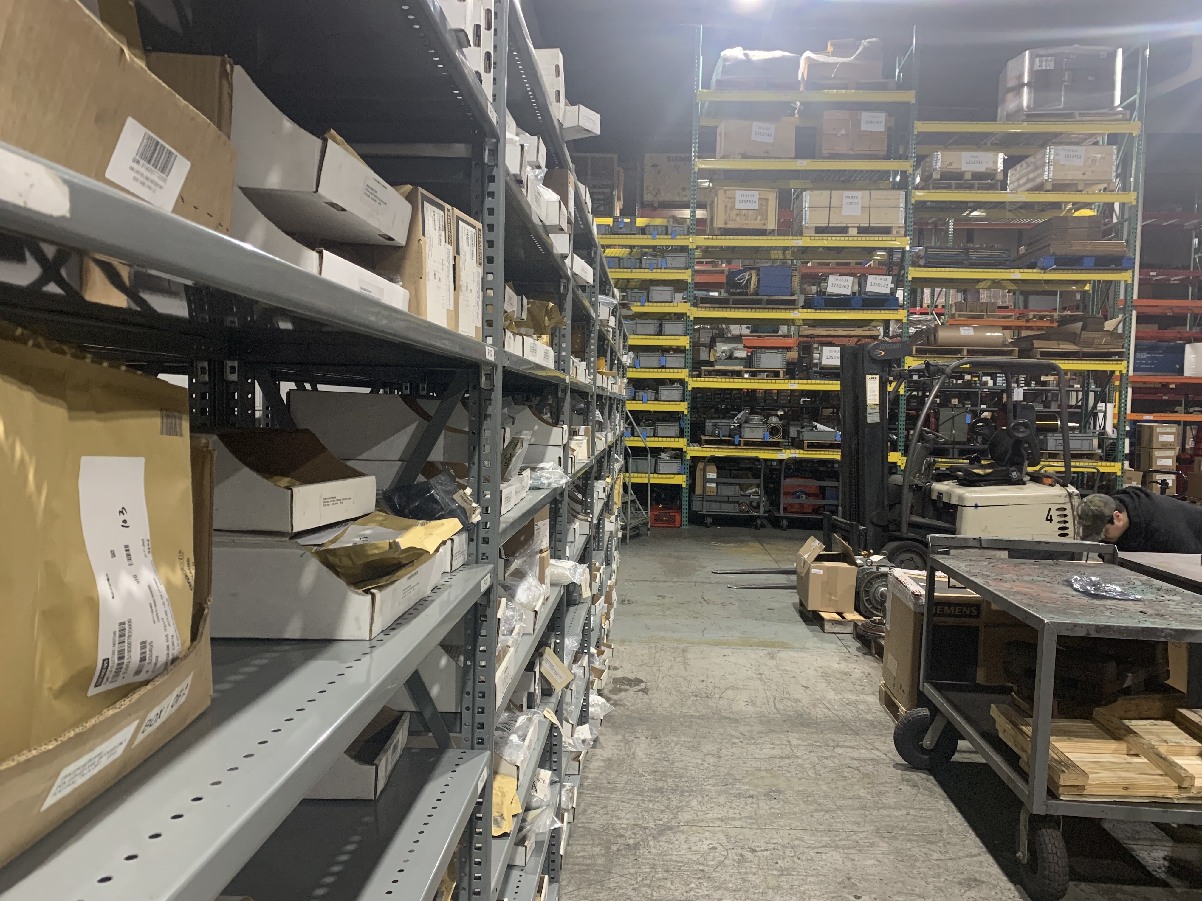 Apex Industrial Automation stock room with close-up of components on shelves and a distant forklift and rolling cart on the right.