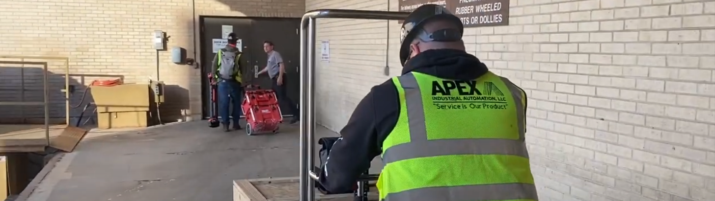 Two Apex Industrial Automation field service technicians entering a customer facility, one being let in by a customer employee, the other pushing a trolley of equipment, both wearing Apex-branded safety gear.