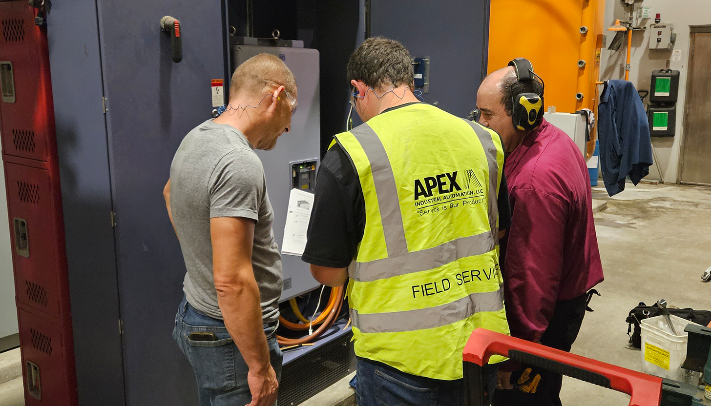 Apex onsite field service engineer explaining retrofitting changes to two customers beside an open control panel in an industrial manufacturing plant.